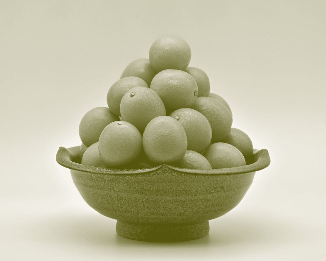 Whole cumquats stacked in a ceramic bowl, their small oval form and glossy skins catching the light.