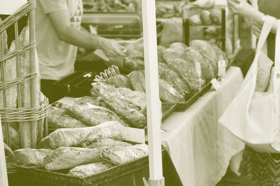 A baker serves customers at a farmers market bread stall stacked with sourdough loaves and baguettes.