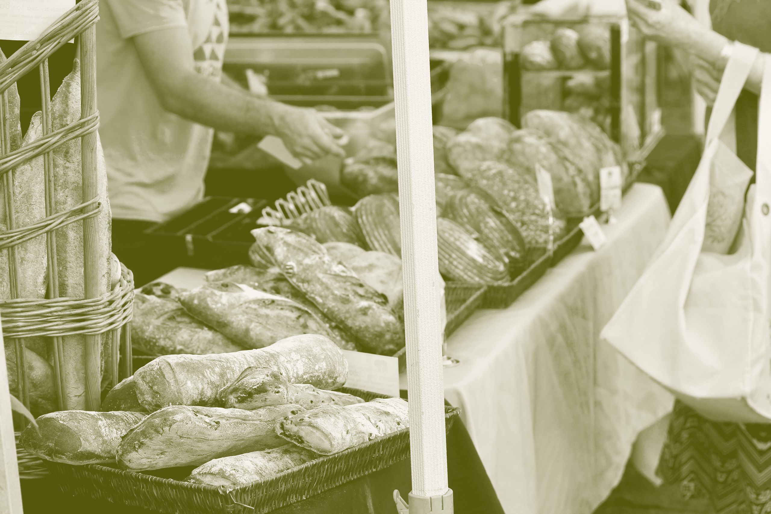 A baker serves customers at a farmers market bread stall stacked with sourdough loaves and baguettes.